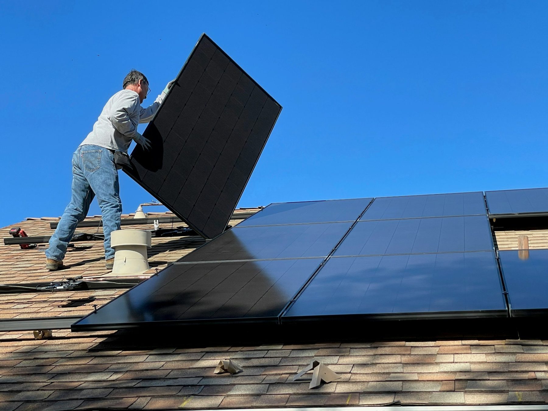 Installer lifting a solar panel into place on a residential roof under a clear blue sky